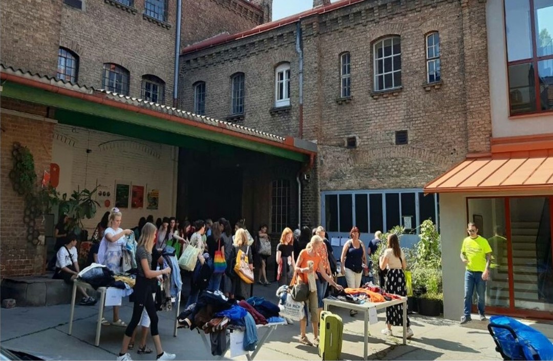 Clothes-swapping event

A colourful mix of people in a Gründerzeit-era inner courtyard, looking at and trying on clothes spread out on tables