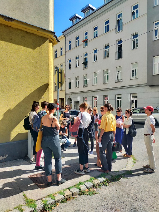 "Inks from Weeds"

A group of young students is seen standing in front of a yellow wall at a street corner, with an older building in the background.
