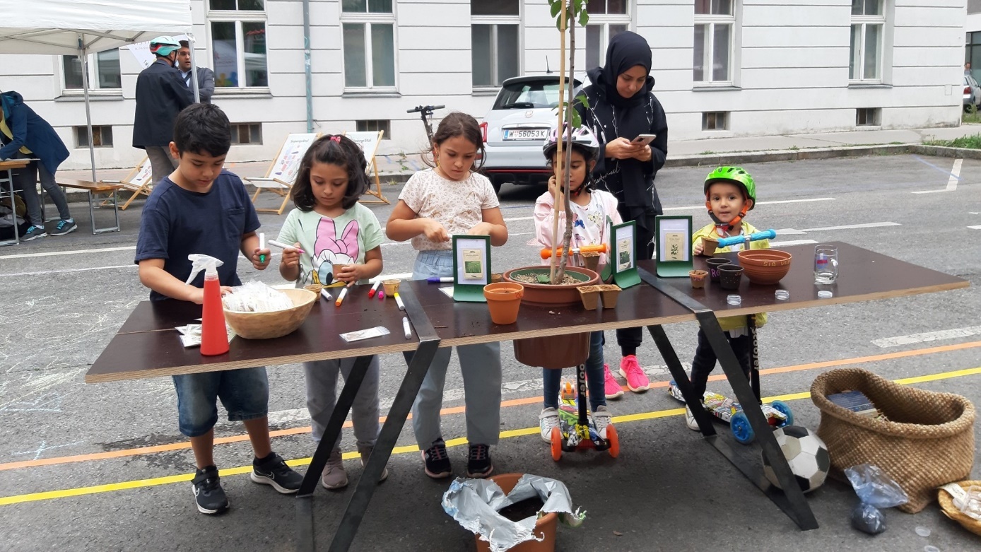 "Grätzlstammtisch"

Five children and a carer are standing at a worktable, planting flowers and labelling the flower pots, with the table put up right on the street.