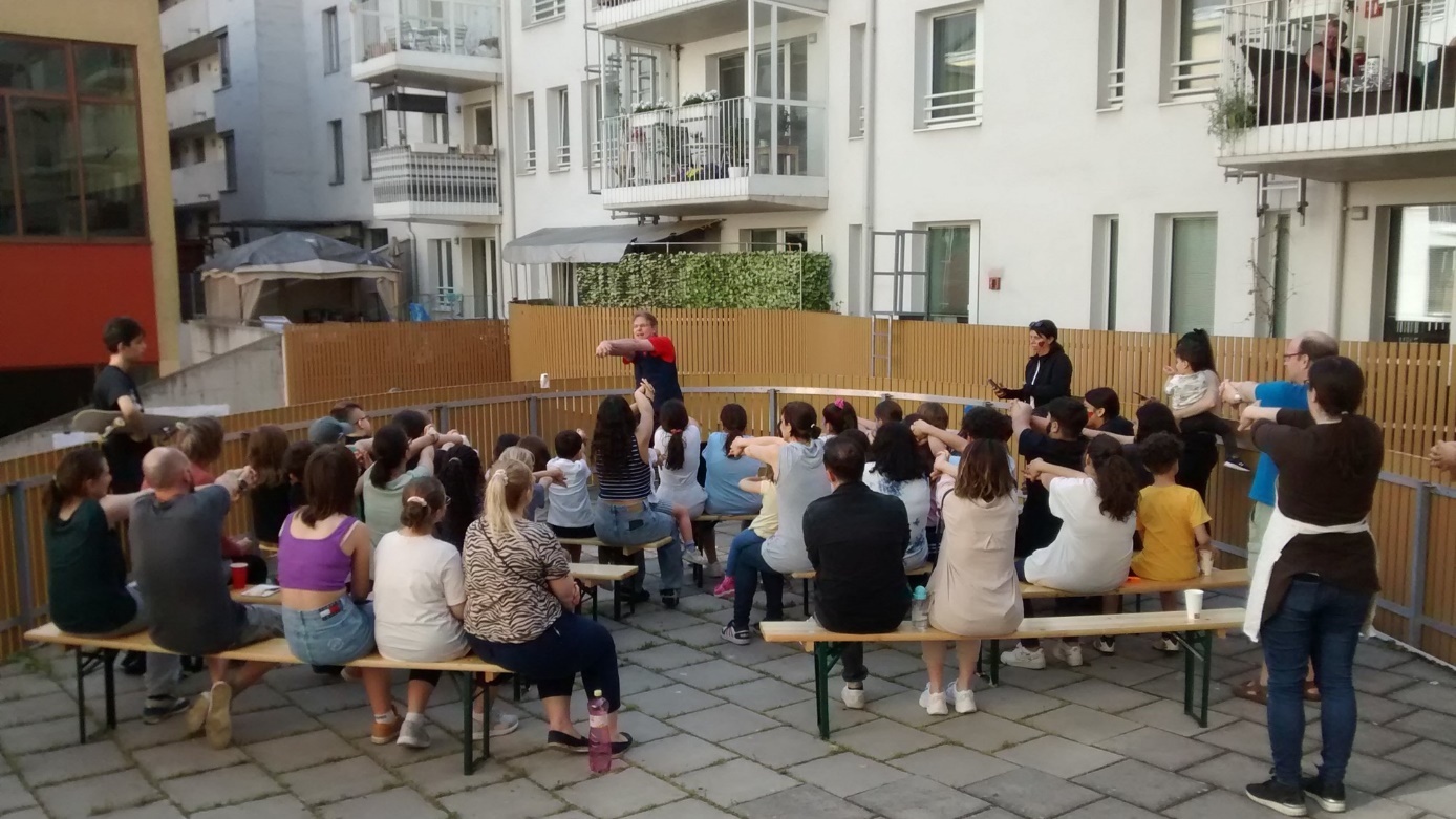 "Miteinander in die Zukunft" (Together Towards the Future)

Spectators are sitting on beer garden benches in the inner courtyard of a residential building and watching a showman's performance. 