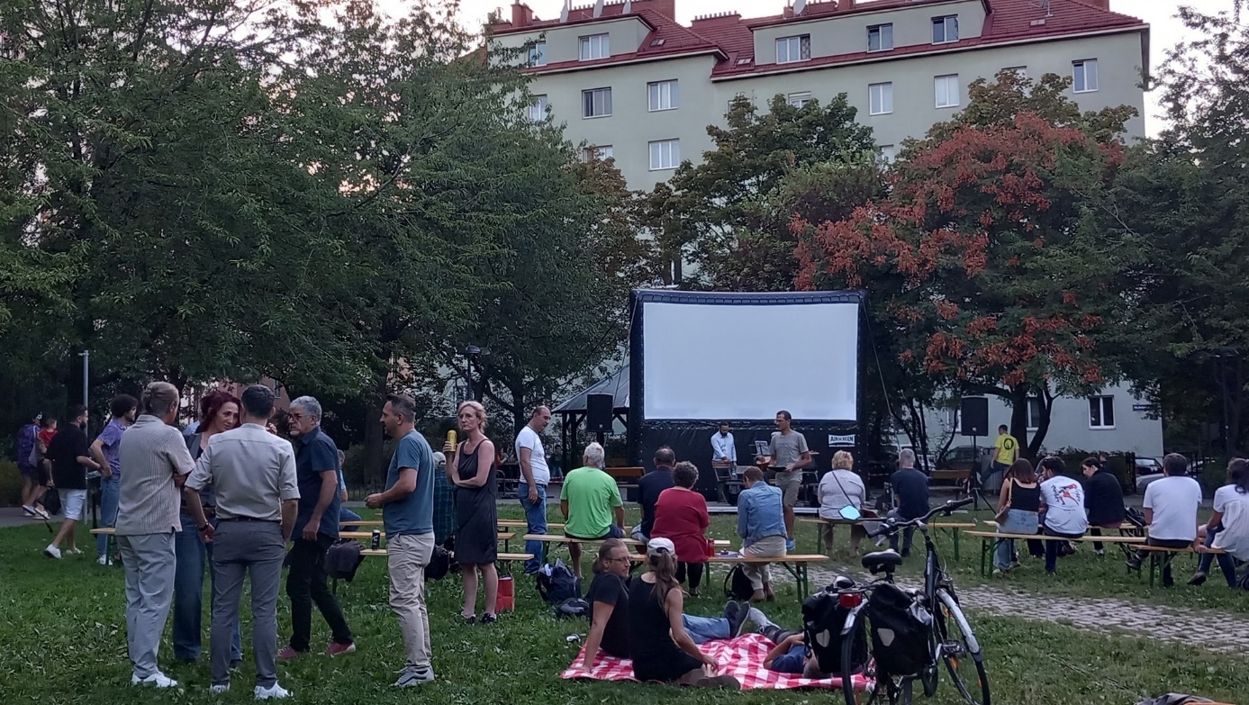 "Grätzlkino"

A white cinema screen in the park, with many people sitting on benches or standing and waiting for the screening to begin