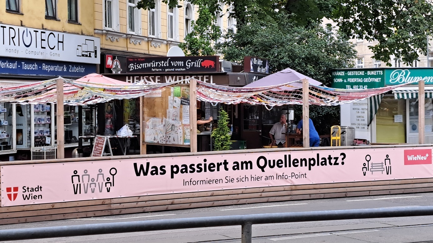 Parklet in Quellenplatz square with text printed on a poster running across the length of the parklet

Parklet with pink poster and lettering in black: "What is happening in Quellenplatz?" 