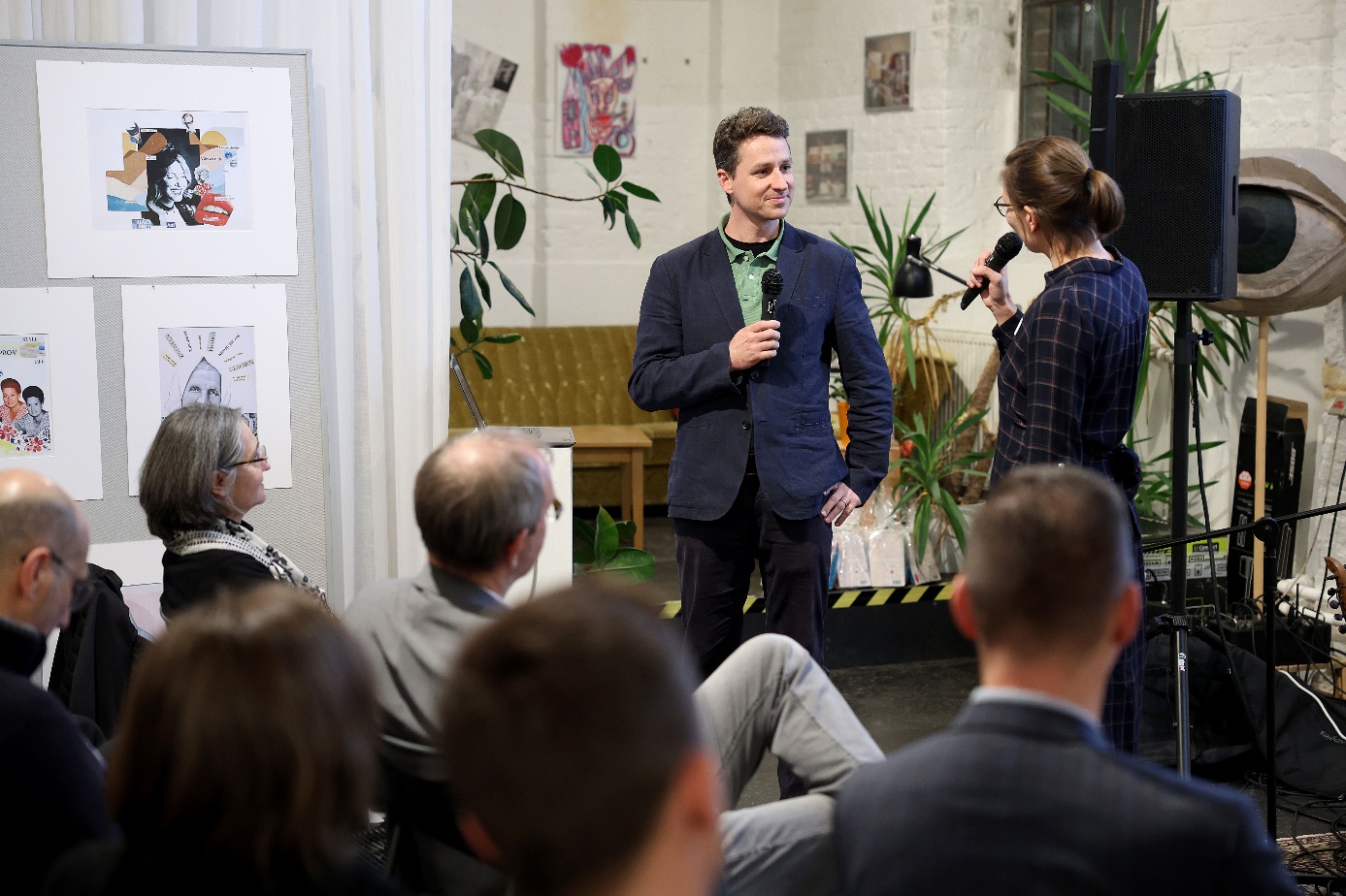 Programme Manager Stephan Hartmann at the final “Grätzlmarie” presentation

Stephan Hartmann is standing in a room before an interested audience, in conversation with a young female participant, with paintings and graphics on the walls as well as plants and various objects advertising the WieNeu+ programme in the background.