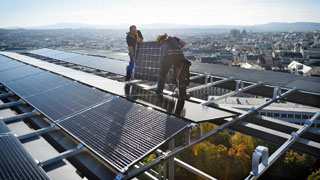 2 Arbeiter montieren Photovoltaik-Paneele auf einem Dach, im Hintergrund Stadtpanorama bei Schnwetter