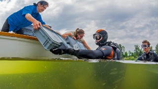 2 auf einem Boot stehende Frauen geben Plastik-Behltnisse an im Wasser befindliche Taucher weiter